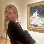 Blond woman standing in an art gallery looking at the camera and smiling.