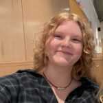 A woman smiling at the camera. She has fair skin, curly blond hair and a blue plaid shirt on.